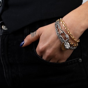 May include: A person wearing a black vest and jeans with a layered bracelet featuring a silver chain, a gold chain, and a silver charm with a diamond accent.