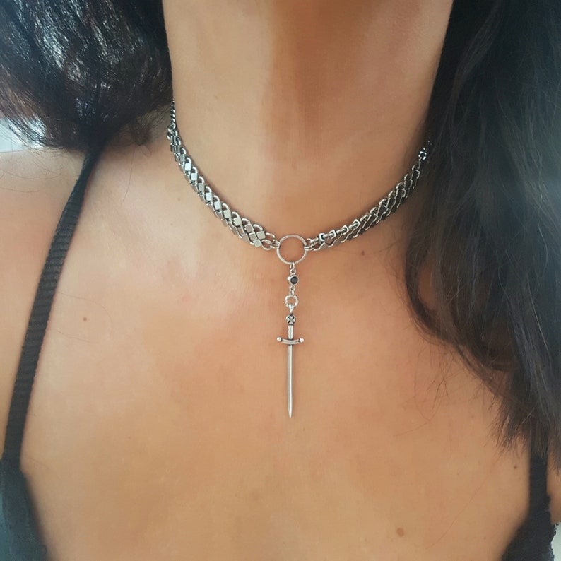 May include: A silver chain choker necklace with a small sword pendant. The chain is thick and has a curb link design. The sword pendant is detailed and has a black stone at the top.