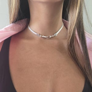May include: A silver choker necklace with a woven design. The necklace has a heart-shaped clasp and decorative end pieces. The jewelry is worn around the neck.