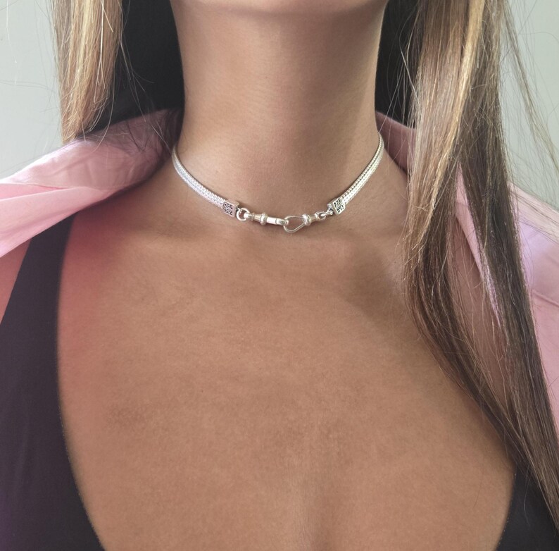 May include: A silver choker necklace with a woven design. The necklace has a heart-shaped clasp and decorative end pieces. The jewelry is worn around the neck.