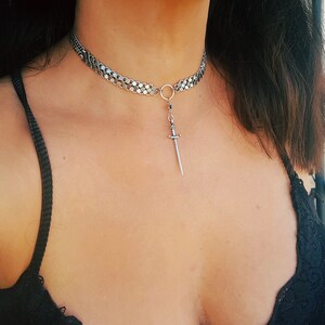 May include: A silver chain choker necklace with a small silver sword pendant. The necklace is worn by a person with dark hair and a black top.