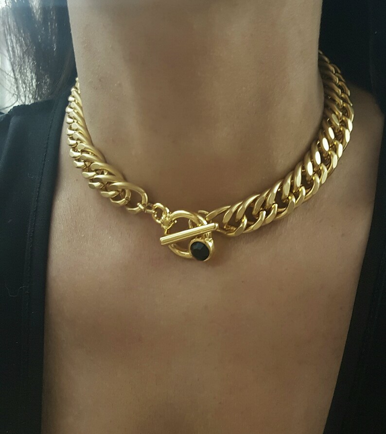 May include: A gold chain necklace with a large, chunky link design. The necklace has a toggle clasp with a black gemstone.