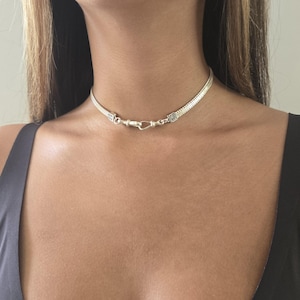 May include: A silver choker necklace with a woven chain design. The necklace features a hook clasp closure and is worn around the neck. The jewelry is a bright silver color and is displayed against a dark background.