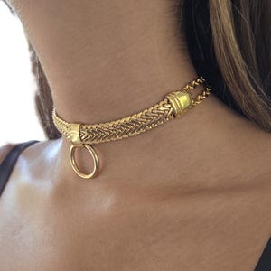 Może przedstawiać: Złoty naszyjnik typu choker z plecionym wzorem i dużym okrągłym wisiorkiem. Naszyjnik wykonany jest z błyszczącego, metalicznego materiału i noszony jest wokół szyi.
