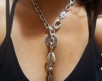 May include: A silver chain necklace with a large coin pendant, a seashell charm, and a tassel. The necklace is layered with a shorter chain with a smaller coin pendant.