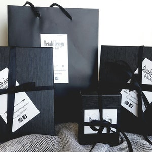May include: Black gift boxes with black ribbon and a white label with the text "Bezalel Design" and contact information. The boxes are on a gray textured surface.