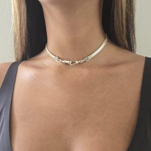 May include: A silver choker necklace with a woven chain design. The necklace features a hook clasp closure and is worn around the neck. The jewelry is a bright silver color.