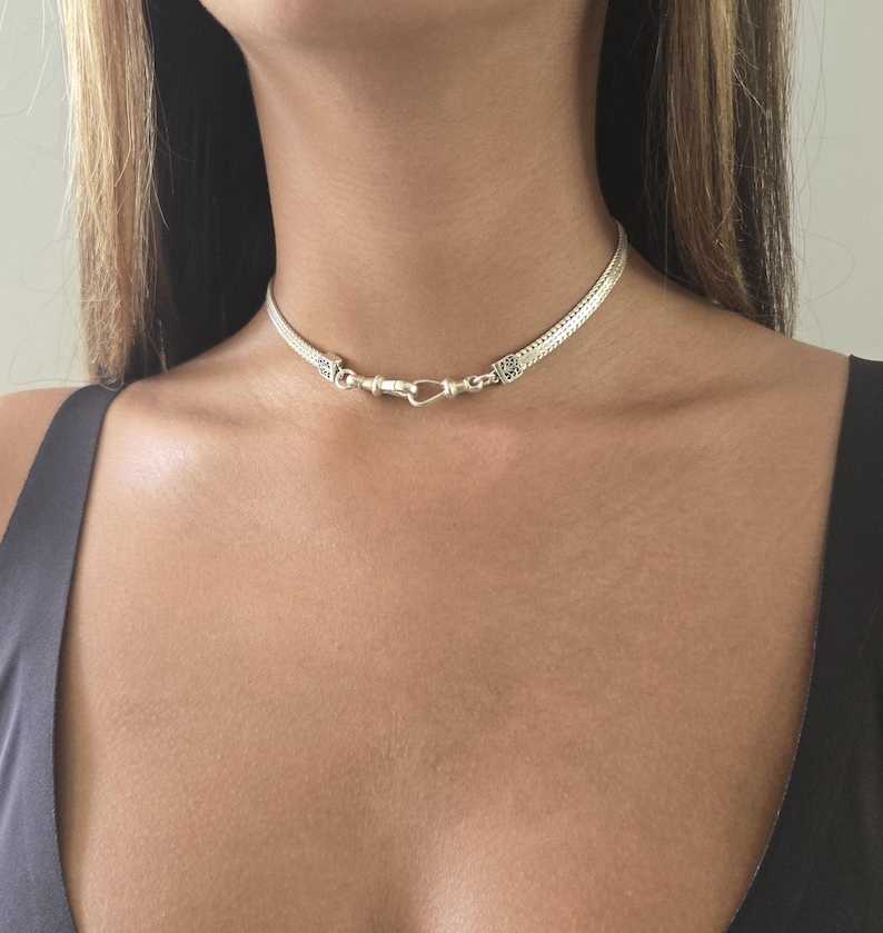 May include: A silver choker necklace with a woven chain design. The necklace features a hook clasp closure and is worn around the neck. The jewelry is a bright silver color.