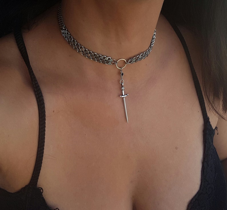 May include: A silver chain choker necklace with a dangling sword pendant. The sword has a black stone at the top.