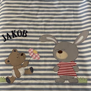 Könnte beinhalten: Ein blaues und weiß gestreiftes Stoff mit dem Namen "JAKOB" in schwarzer Stickerei. Ein brauner Teddybär, der eine rosa Blume hält, und ein graues Kaninchen, das ein rot-weiß gestreiftes Hemd trägt, sind auf den Stoff appliziert.