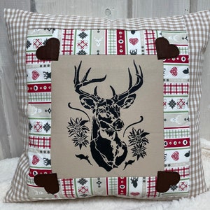 Puede incluir: Cojín decorativo cuadrado con un motivo de cabeza de ciervo negro sobre fondo beige. El cojín tiene un borde estampado con detalles en rojo, verde y beige, y detalles en forma de corazón marrón en las esquinas. El borde exterior es un estampado de cuadros vichy beige y blanco.