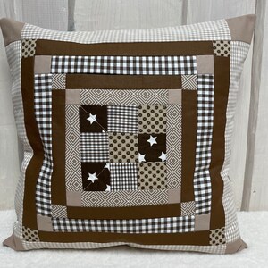Peut inclure: Un coussin patchwork marron et beige avec un motif carré au centre. Le carré central est composé de carrés plus petits dans différents motifs, notamment du vichy, des pois et des motifs géométriques. Il y a des étoiles blanches sur certains des carrés.