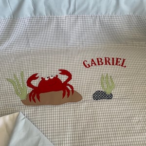 Op de afbeelding: Een grijs en wit gingham wiegbeddengoedset met een rode krab-applicatie en de naam "GABRIEL" geborduurd in rood.