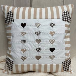 May include: A beige and white striped cushion with a white centre panel featuring a quilted design of black and brown hearts. The cushion has a brown and white gingham border.