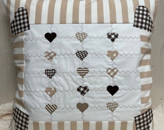 May include: A beige and white striped pillow with a white center panel featuring a quilted design of black and brown hearts. The pillow has a brown and white gingham border.