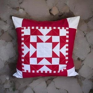 Könnte beinhalten: Quadratisches Zierkissen mit einem geometrischen Sternmuster in Rot und Weiß. Das Kissen hat einen roten Hintergrund mit weißen Stern- und Quadratformen. Die Ecken sind weiß.