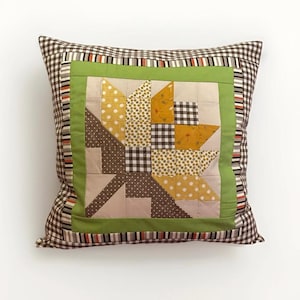 May include: Square decorative pillow with a patchwork design. The pillow features a central floral pattern in shades of yellow, brown, and cream, framed by a green border and a brown and white checkered pattern. The pillow is a square shape.