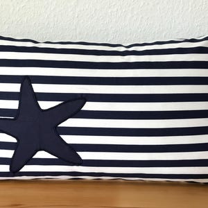 Könnte beinhalten: Rechteckiges Kissen mit marineblauen und weißen horizontalen Streifen. Auf der Vorderseite befindet sich ein Applikation in Form eines dunkelblauen Seesterns. Das Kissen hat ein Seildetail an einem Ende. Nautisches Thema.