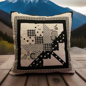 Puede incluir: Cojín decorativo cuadrado con un diseño de patchwork en blanco y negro. El cojín presenta varios patrones, incluyendo lunares, cuadros vichy y motivos florales. Un borde negro y un borde de cuadros vichy enmarcan el diseño. El cojín está sobre una superficie de madera.
