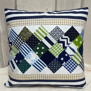 Op de afbeelding: Een wit kussen met een blauw en groen patchwork ontwerp. Het kussen heeft een blauwe en witte gestreepte rand en een beige ruitjesrand.
