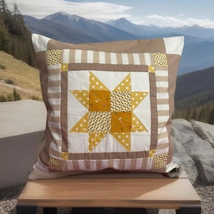 May include: A square cushion with a star quilt design. The cushion features a central star pattern in yellow and white, surrounded by a brown border and striped accents. The cushion is placed on a wooden surface with a mountain backdrop.