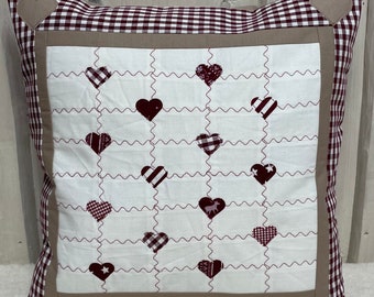 May include: A white pillow with a brown and white gingham border. The pillow has a brown frame with a white background and a pattern of red hearts with different designs inside each heart.