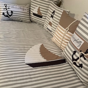 Könnte beinhalten: Eine gestreifte graue und weiße Bettdecke mit dekorativen Kissen. Die Kissen zeigen nautische Motive, darunter Anker, Segelboote und Leuchttürme. Die Kissen tragen den Text "Heimathafen" und "Liebplatz".