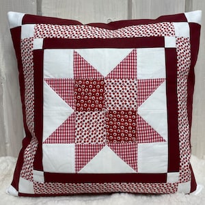 Könnte beinhalten: Quadratisches Kissen mit einem Sternmuster aus Patchwork. Das Kissen hat einen bordeauxfarbenen Hintergrund mit einem weißen und bordeauxfarbenen Sternmuster in der Mitte. Der Stern ist von weiß-roten Vichy- und Blumenmustern umgeben.