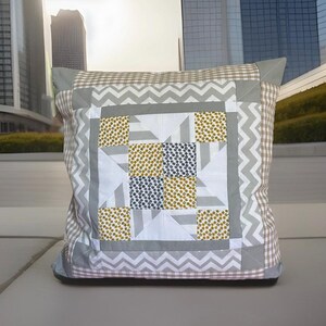 Peut inclure: Coussin décoratif carré avec un motif patchwork. Le coussin présente un carré central avec des motifs floraux jaunes et marine, entouré de formes géométriques grises et blanches. La bordure a un motif en zigzag gris et un motif vichy marron et blanc.