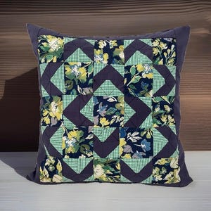 Peut inclure: Un coussin carré au design patchwork avec une bordure violet foncé. Le motif central se compose de blocs alternés de tissu bleu marine foncé avec des imprimés floraux jaunes et blancs, et de carrés de tissu à carreaux vert clair et blanc.
