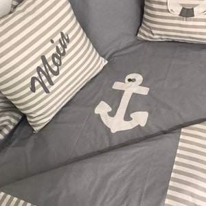 Könnte beinhalten: Eine graue und weiße gestreifte Decke mit einem weißen Anker-Design und einem Kissen mit dem gestickten Wort "Moin".
