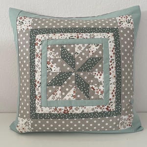 Puede incluir: Cojín decorativo cuadrado con diseño de patchwork. El cojín presenta un motivo de estrella central en gris y blanco, rodeado de bordes estampados en gris, blanco y estampados florales. El borde exterior es azul claro liso.