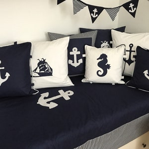 Könnte beinhalten: Eine dunkelblaue Couch mit weiß-blau gestreiftem Besatz. Die Couch ist mit Kissen mit nautischen Motiven bedeckt, darunter Anker, ein Schiff und ein Seepferdchen. Die Kissen sind weiß mit dunkelblauen Motiven.
