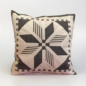 Può includere: Cuscino quadrato decorativo con un design geometrico a stella. Il cuscino presenta uno sfondo beige chiaro con motivi bianchi e neri, tra cui strisce e pois. I bordi sono rifiniti con un bordo grigio scuro.