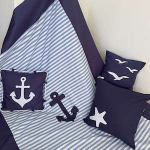 Può includere: Una coperta a righe blu e bianche con un bordo blu scuro. La coperta ha due cuscini con ancore bianche e un cuscino con una stella marina bianca. La coperta è perfetta per una camera da letto a tema nautico.