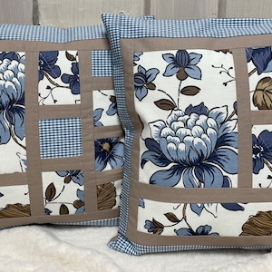 Könnte beinhalten: Zwei dekorative Kissen mit Patchwork-Muster. Die Kissen bestehen aus weißem Stoff mit blauen und braunen Blumenmustern. Der Stoff ist in einem Gittermuster zusammengenäht, wobei brauner Stoff jedes Quadrat umrahmt.