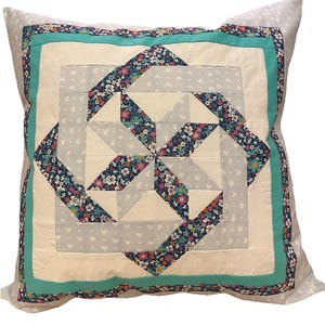 Può includere: Un cuscino decorativo con sfondo bianco e bordo turchese. Il centro del cuscino presenta un motivo geometrico di tessuto floreale in tonalità di blu, rosa e bianco.