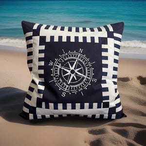 Könnte beinhalten: Quadratisches Zierkissen mit nautischem Thema. Es hat eine marineblaue Mitte mit einem weißen Kompassdesign. Der Rand ist marineblau und weiß gestreift. Das Kissen liegt an einem Sandstrand mit dem Meer im Hintergrund.