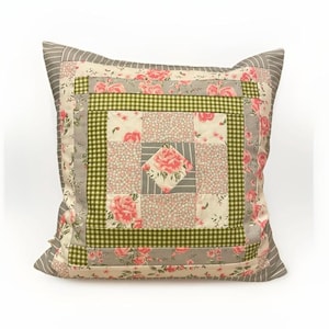 Può includere: Cuscino quadrato decorativo con un design patchwork. Il cuscino presenta un quadrato centrale con un motivo floreale, circondato da quadrati di tessuti floreali, a quadretti e a righe. La palette di colori comprende rosa, verde, grigio e crema.