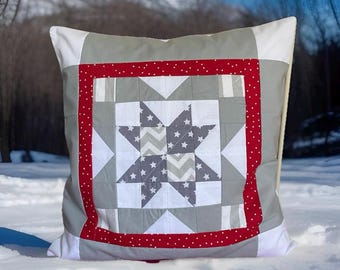May include: Square decorative pillow with a patchwork design. The pillow features a central star pattern in gray and white, framed by a red border with white polka dots. The outer edges are gray and white.