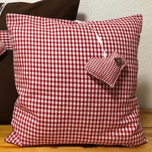Könnte beinhalten: Ein Kissen mit rot-weißem Vichy-Muster, verziert mit einem herzförmigen Akzent. Das Herz ist aus dem gleichen Stoff gefertigt und hat einen kleinen Holzknopf. Im Hintergrund ist ein braunes Kissen teilweise zu sehen.