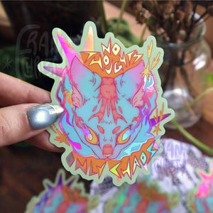 May include: A holographic sticker depicting a cat-like creature with a unicorn horn, in shades of blue, pink, and orange. The text "NO THOUGHTS ONLY CHAOS" is written around the cat's head. The sticker has a light green border.