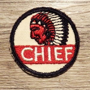 May include: A round, white embroidered patch with a black outline. The patch features a red and black image of an American Indian chief wearing a feathered headdress. The word "CHIEF" is embroidered in white on a red banner below the image.