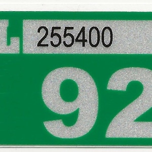 May include: A rectangular sticker with a green background and white border. The sticker features the letter "L" and the number "255400" in a silver rectangle, and the number "92" in large silver digits.