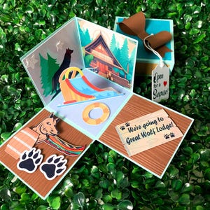 May include: A handmade pop-up card with a Great Wolf Lodge theme. The card features illustrations of a water slide, a cabin, and a wolf. Text on the card reads "Open for a Surprise!" and "We're going to Great Wolf Lodge!"