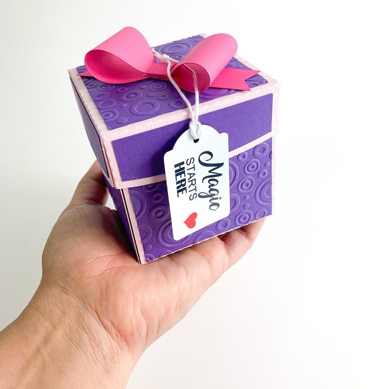 May include: A small purple gift box with a pink bow and a white tag that says "Magic Starts Here" with a red heart.