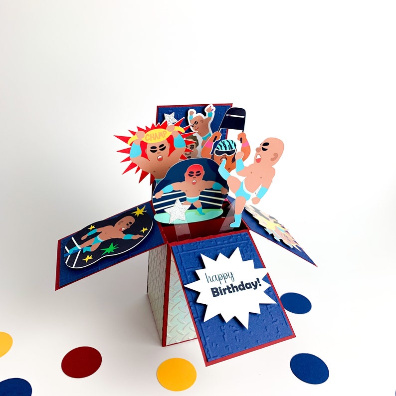 Puede incluir: Una tarjeta emergente azul y roja con tem&aacute;tica de lucha libre. La tarjeta presenta un ring de lucha libre con luchadores en acci&oacute;n. La tarjeta est&aacute; abierta y el interior dice "&iexcl;Feliz cumplea&ntilde;os!"