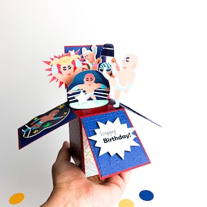 Puede incluir: Una tarjeta de cumplea&ntilde;os pop-up con tem&aacute;tica de lucha libre. La tarjeta est&aacute; abierta y muestra un ring de lucha libre con cuatro luchadores en acci&oacute;n. La tarjeta tiene un fondo azul y rojo con una estrella blanca en el centro. La tarjeta tambi&eacute;n tiene una estrella blanca con el texto "happy Birthday!" en negro.
