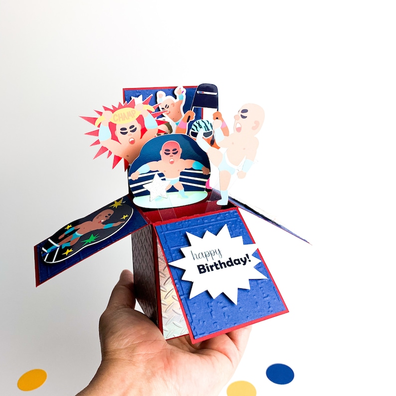 Puede incluir: Una tarjeta de cumplea&ntilde;os pop-up con tem&aacute;tica de lucha libre. La tarjeta est&aacute; abierta y muestra un ring de lucha libre con cuatro luchadores en acci&oacute;n. La tarjeta tiene un fondo azul y rojo con una estrella blanca en el centro. La tarjeta tambi&eacute;n tiene una estrella blanca con el texto "happy Birthday!" en negro.