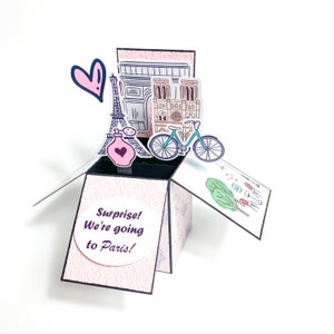 May include: A pink and black pop-up card with a surprise message that reads "Surprise! We're going to Paris!" The card features a pop-up Eiffel Tower, Notre Dame Cathedral, the Arc de Triomphe, a bicycle, and a bottle of perfume.